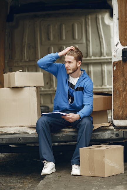 A young man with light brown hair and a beard, wearing a blue jacket over a white shirt, sits inside the open rear of a moving van, surrounded by cardboard boxes. He holds a clipboard in his left hand