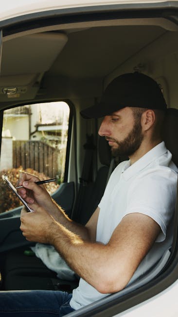 A man with glasses, wearing a light blue shirt and dark trousers, is seated on a wooden chair next to a white moving van parked on the street outside a modern building with glass windows and a white f