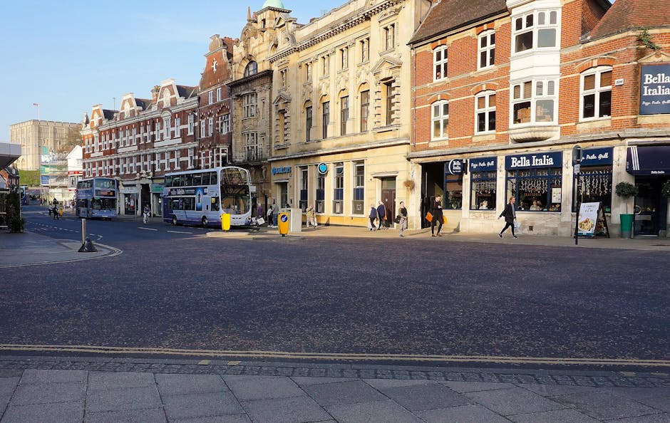 A street scene on Sutton High Street featuring multiple multi-storey buildings with mixed architectural styles, including red brick and ornate stone facades, housing various shops and businesses. Two double-decker buses are parked along the pavement near the curb, with some passengers visible inside. Several pedestrians are walking along the sidewalk, some carrying bags or backpacks, and others standing near the entrance of a restaurant called Bella Italia, which has a blue awning and outdoor seating area. The street is paved with asphalt, with visible road markings and a yellow double line along the curb. Lighting suggests it is daytime with clear skies, and the overall environment depicts an active urban area, ideal for house removals or moving services as handled by Sutton Movers, who operate in this locality. This scene captures a typical busy high street, highlighting the logistics and infrastructure involved in furniture transport and home relocation services in Sutton.