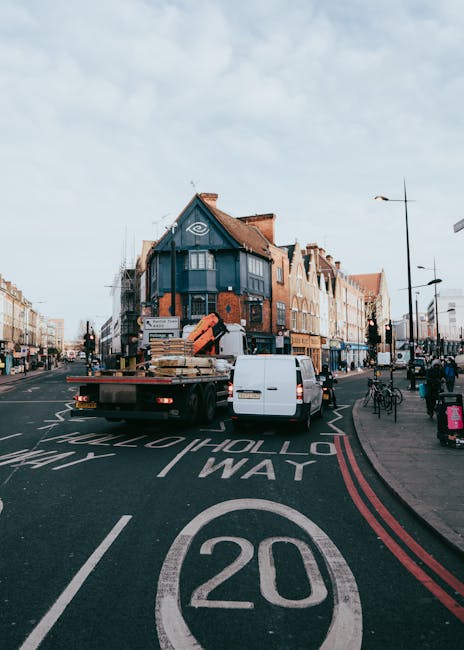 A street scene on Sutton High Street during a home relocation process, showing a flatbed truck with a low-loading platform parked on the road, loaded with various furniture items including large cardboard boxes, wooden pallets, and wrapped objects. The truck is positioned near the curb, with a white van parked behind it, likely part of the furniture transport. The street features typical urban shopfronts and buildings, with pedestrians walking along the sidewalk and bicycles parked nearby. The road markings include a speed limit of 20 miles per hour and directional arrows, indicating it is a busy shopping area within Sutton. Overhead, the sky is partly cloudy with daylight illuminating the scene, capturing the moving logistics involved in house removals as part of a professional relocation service by Sutton Movers. The presence of moving equipment such as blankets, straps, or trolleys is not visible, but the scene suggests active packing and loading for a house removal or furnishings transportation within the area.
